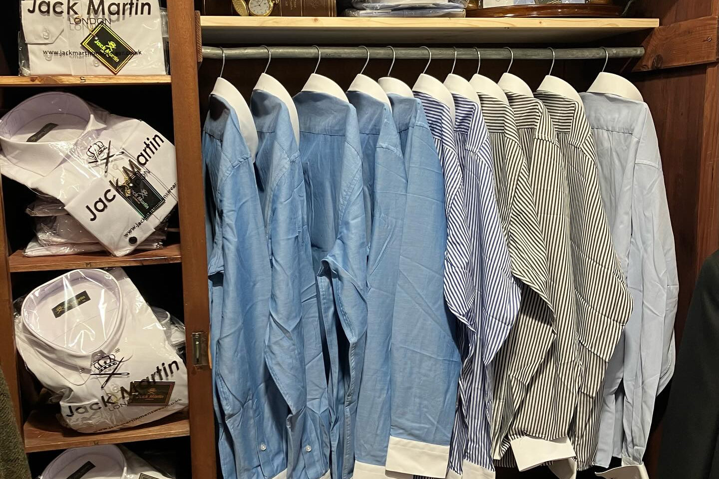 A wooden closet organizer filled with Jack Martin dress shirts, including folded white shirts on the left shelves, a row of light blue and striped shirts hanging in the center, and more shirts and boxes on the lower shelf.