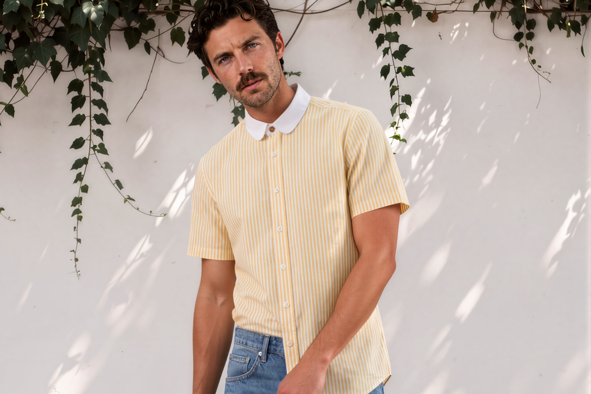 A man wearing a pale yellow ribbed short-sleeve button-up shirt with a white collar, blue denim shorts, and brown sandals, standing against a white wall with leafy vines.